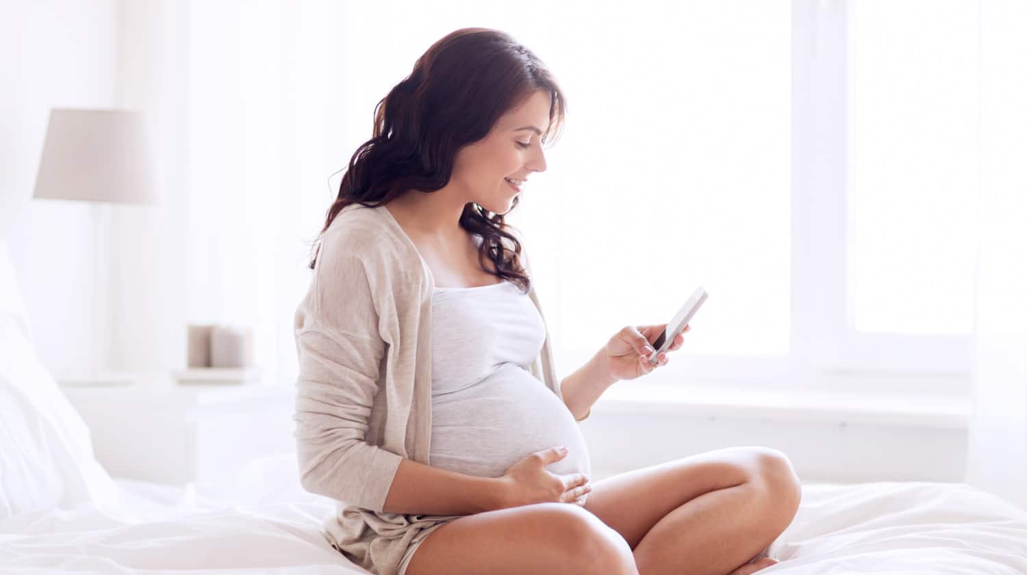 Happy pregnant woman with smartphone in bed | Most Helpful Pregnancy Apps | glow pregnancy app | Featured