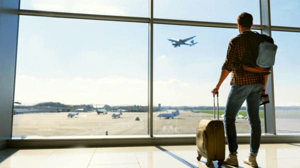 Feature | Young man is standing near window at the airport and watching plane before departure | Travel Tips For The Newbie International Traveler